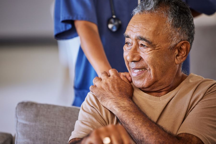 A smiling older man, with dark skin tone, sitting in a sofa chair. A person in blue scrubs behind him holding his shoulder